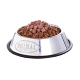 A stainless steel pet food bowl, filled with dry dog kibble and topped with bite-sized morsels of Primal Butcher's Blend raw meal topper.