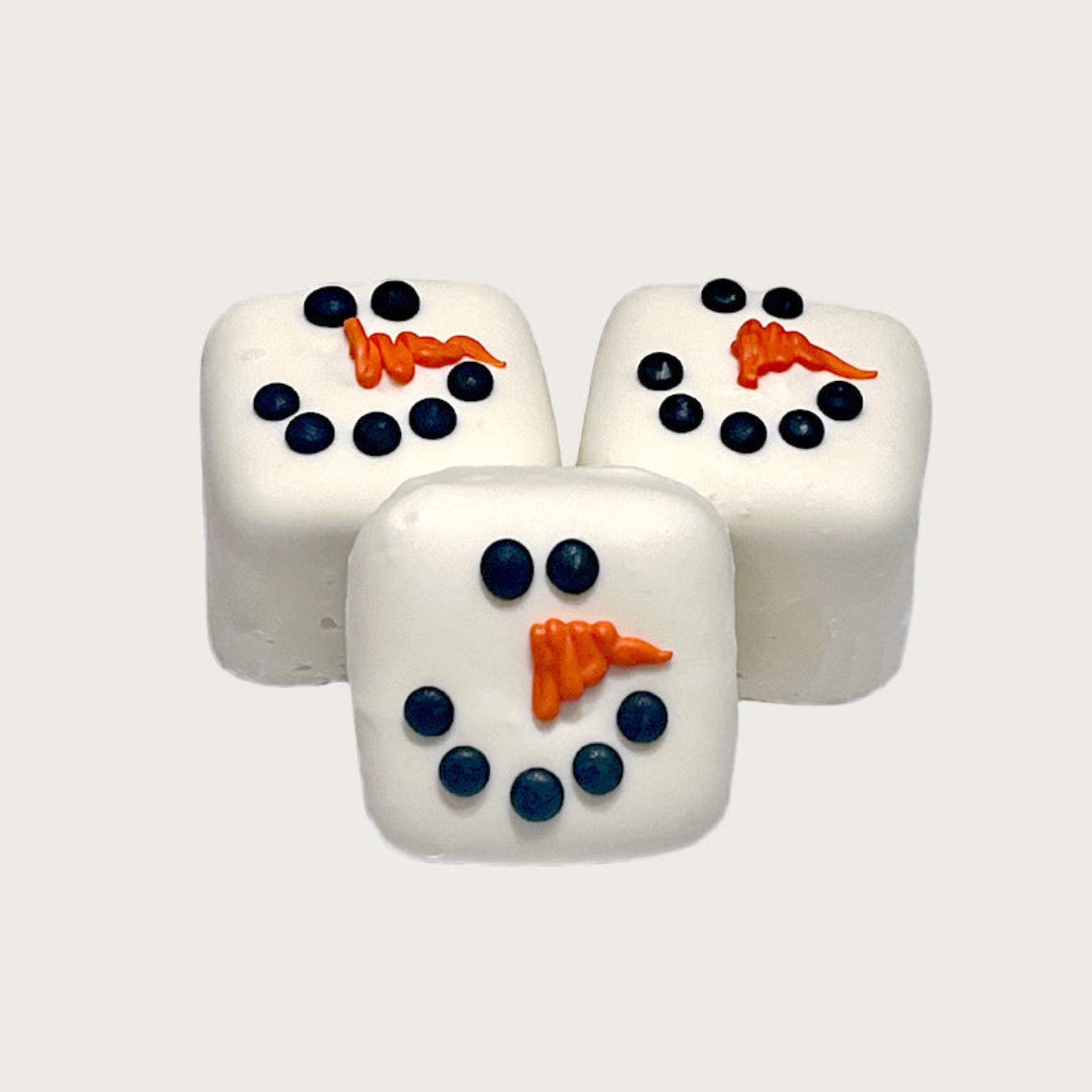 Three square Bon Bon dog treats, decorated to look like snowmen. They are white with black dots for eyes & mouth, and have an orange frosted nose in the shape of a carrot. 