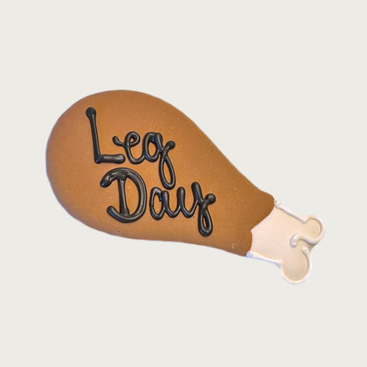 Decorated dog cookie in the shape of a cooked turkey leg (drumstick). Brown "leg" with white tip, and "Leg Day" written in dark brown script text.