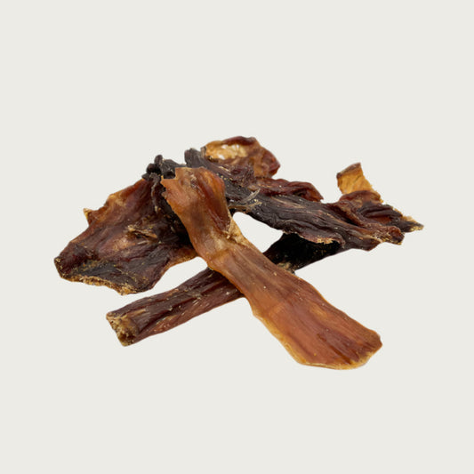 Closeup photo of a small pile of Wet Nose Road Runner Stix dog treats. Product is varying sizes of rectangular shaped, thin brown strips, ranging from approximately 3" - 6" long.